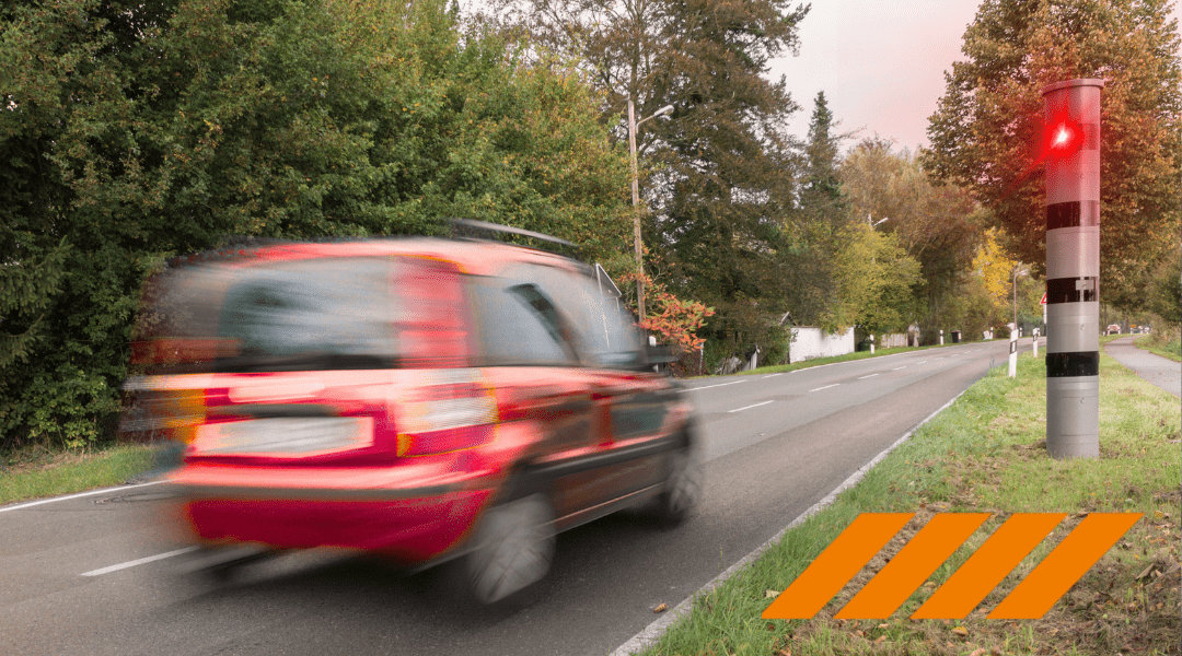 Blurry red car speeds along a two-lane road with trees on both sides; a striped post with a red signal stands on the right, and orange diagonal hazard markings are in the foreground.